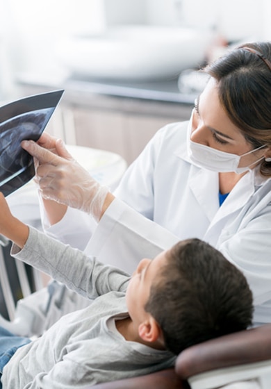 Uma dentista de jaleco branco e máscara facial mostrando um raio-X dentário a um menino sentado em uma cadeira odontológica.