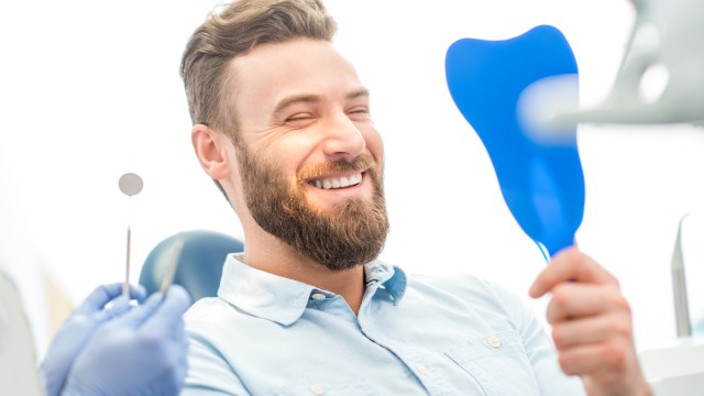 Homem sorridente sentado na cadeira de um dentista, vendo o seu sorriso através de um espelho.